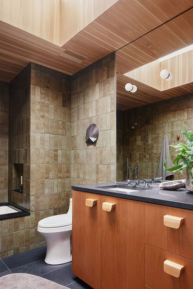 A richly detailed bathroom with a warm mid-century character. The room features dark brown and mottled beige tiled walls, a dark tiled floor, a timber vanity with chunky cream-colored drawer pulls, a dark benchtop, a white basin and toilet, and a large mirrored wall. The ceiling is clad in warm timber panels with recessed geometric sections and round white light fixtures. Plants and sculptural accessories add natural accents.