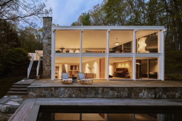 A modern two-storey glass-fronted house set into a wooded landscape, photographed at dusk. The building features extensive floor-to-ceiling glazing, slender light-coloured structural framing, a prominent rough natural-stone chimney and retaining wall, timber decking, external stairs, and a reflecting pool or water feature in the foreground. Warm interior lighting reveals living and dining spaces inside.