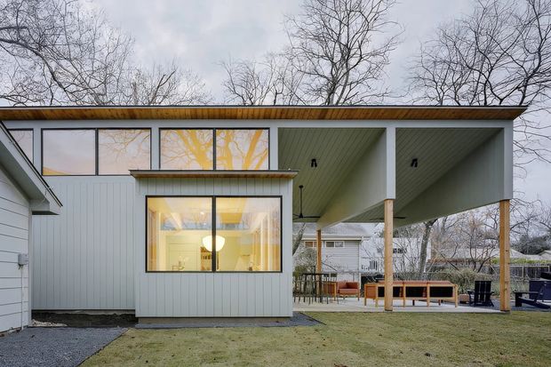 Exterior view of a contemporary single-storey house extension with pale vertical cladding, broad flat roof, clerestory-style glazing, and a large covered outdoor area supported by timber posts. Warm interior lighting is visible through the windows, with a furnished patio beneath the roof overhang. Leafless trees and neighbouring residential structures form the background.