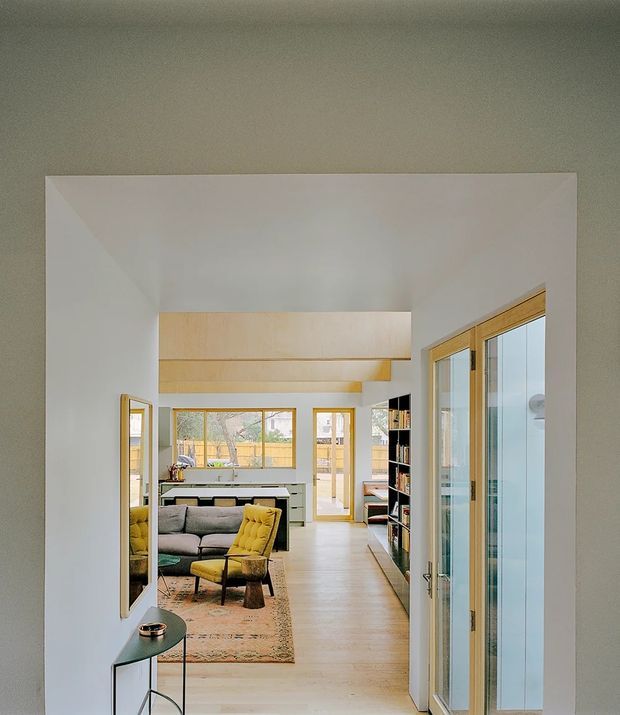 Bright contemporary open-plan interior viewed from a hallway or entry passage, looking toward a living room and kitchen. The space has white walls and ceilings, pale timber flooring and built-in cabinetry, extensive timber-framed glazing and sliding doors, a grey sofa, mustard lounge chair, patterned rug, and black open shelving. A high ceiling and strong natural light create an airy, connected layout.