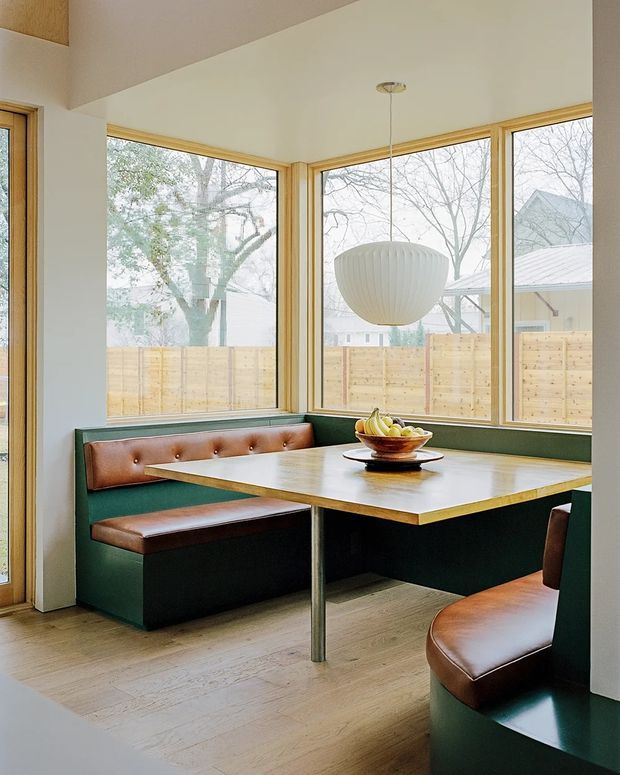 Bright dining nook with a built-in L-shaped green banquette, brown leather seat cushions, and a rectangular wood-topped table with a central metal pedestal. Floor-to-ceiling corner windows provide abundant natural light, while a large white pendant hangs above the table. The room has pale timber flooring, white walls, warm timber window frames, and a view of a fenced yard and neighboring houses outside.