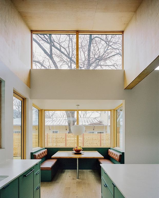 Bright contemporary dining nook viewed from a kitchen, with built-in green cabinetry and white benchtops in the foreground. The dining area has a central table, leather-upholstered banquette seating, a sculptural pendant light, and extensive timber-framed glazing. A high clerestory window and elevated ceiling create a strong sense of vertical volume, while pale timber lining continues across the ceiling and upper walls.