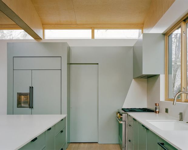 A bright, compact contemporary kitchen with pale sage-green cabinetry, white benchtops, integrated appliances, and a deep single-bowl sink beneath timber-framed windows. The room has a distinctive plywood-lined ceiling and clerestory glazing that brings in abundant natural light. A central island occupies the foreground, while the cooking zone and tall integrated refrigerator cabinetry line the opposite wall.