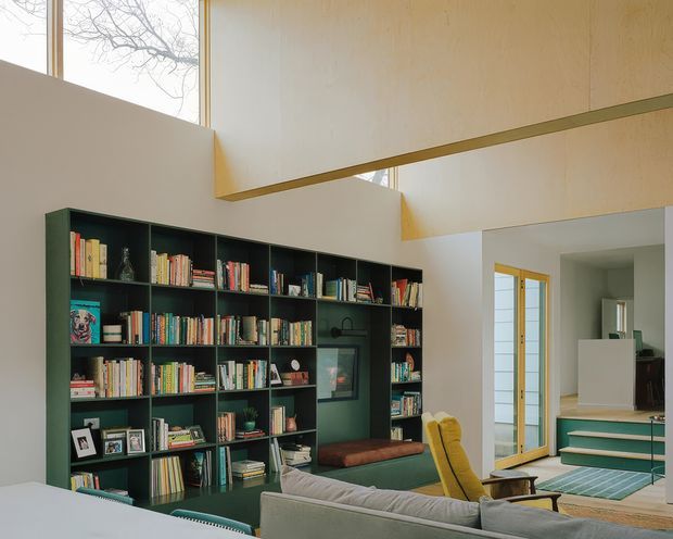 A bright, double-height living room dominated by a full-height dark green built-in bookshelf filled with books and objects. A built-in window seat, grey sofa, mustard lounge chair, and timber-framed glazing create a warm, eclectic reading and living area. Pale timber or plywood-clad overhead architectural volumes project across the upper space, while teal carpeted steps lead to an adjoining level. High windows bring in natural light and overlook bare tree branches.