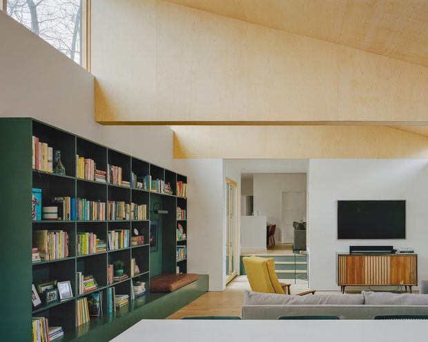 Bright contemporary living room with a large dark green built-in bookshelf filled with books, a timber floor, pale timber-lined ceiling and upper wall, and a neutral sofa facing a wall-mounted television. A mustard lounge chair and timber media console add warm accents. The open doorway reveals a secondary interior area beyond.