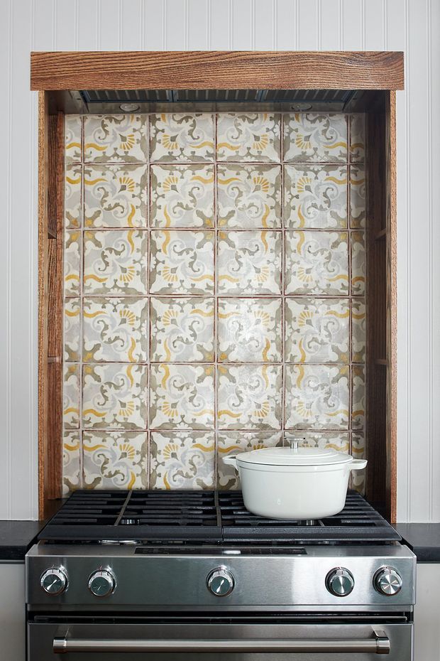 Close-up view of a freestanding stainless-steel range cooker set into a dark timber-framed alcove. The alcove has a decorative patterned ceramic tile splashback in muted cream, grey, beige and mustard tones, with white vertical wall panelling surrounding it. A white lidded cookware pot sits on the cooktop.