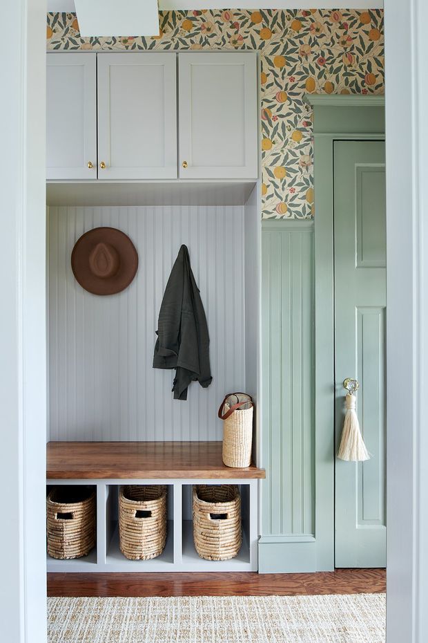 A compact, carefully styled entryway with pale sage-green and white built-in cabinetry, a warm timber bench seat, open cubbies containing woven baskets, beadboard wall panelling, and hooks for coats and a hat. Botanical and citrus-patterned wallpaper adds a colourful decorative feature, while a paneled sage-green door with a tassel handle is visible at right. A woven rug and natural fibre accessories contribute to a warm, traditional cottage-like atmosphere.