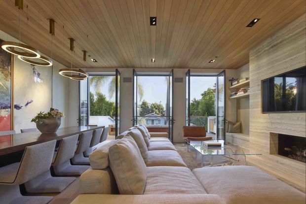 Open-plan living and dining room with a large upholstered sectional sofa facing a built-in fireplace and television wall. The room features extensive floor-to-ceiling glazing with operable panels, a warm timber-lined ceiling, timber or stone-look wall finishes, contemporary ring pendant lights, and built-in shelving. A dining table occupies the left side, while leafy residential views are visible through the windows.
