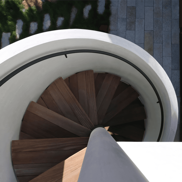 Looking down through a compact curved stairwell, with dark timber spiral stair treads winding around a central smooth white structural column. The stair is enclosed by a sculptural white circular wall and slim black metal handrail, with grey stone paving visible beyond the opening at the top. Strong geometric composition, contrasting white plaster, dark timber, and black metal, photographed from above.