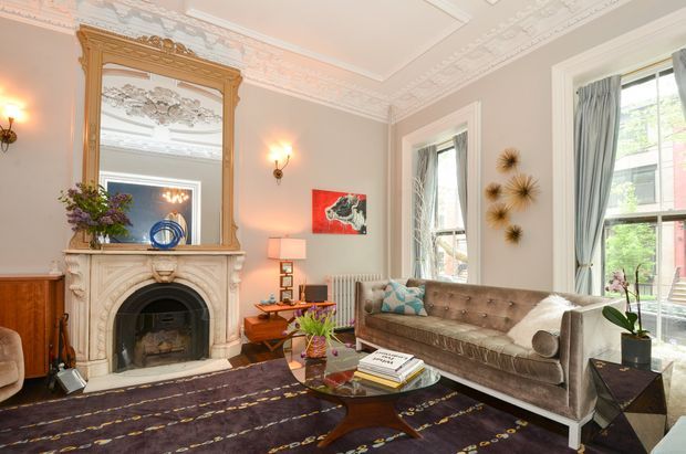 Elegant traditional living room with ornate plaster ceiling mouldings, a decorative carved fireplace and overmantel mirror, tall sash windows with drapery, and a tufted sofa arranged around a sculptural wooden coffee table. The room combines warm neutral walls with dark patterned flooring and richly detailed period features.