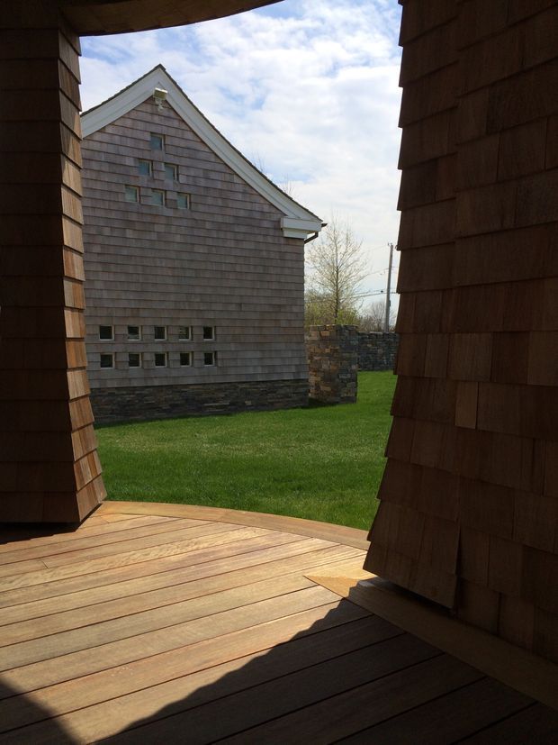 View from a timber-clad outdoor deck or covered terrace framed by dark cedar-shingle walls, looking across a lawn toward a separate gable-roofed building. The background building has weathered shingle cladding, white gable trim, small square windows, and a dark stone base, with a low stone wall beyond.