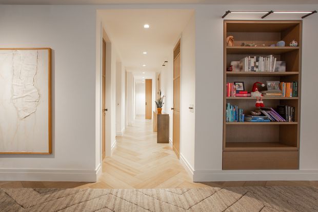 A refined residential hallway viewed from a living area, with pale herringbone timber flooring, white walls and trims, recessed ceiling lights, and multiple doorways leading down the corridor. A built-in dark timber bookcase with books and decorative objects occupies the right wall, while a framed neutral artwork hangs on the left. The space has a warm, calm and contemporary feel.