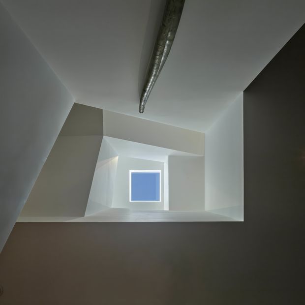 A minimalist architectural view looking upward through a geometric stairwell or vertical void. Angular white and light-grey walls form a sharply faceted composition around a small square blue window, while an exposed metallic duct or structural element runs along the ceiling. The image has a subdued, sculptural, high-contrast atmosphere.
