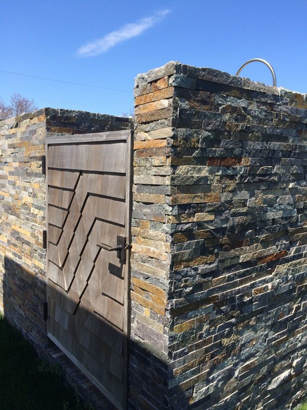 Exterior detail of a low, heavily textured dry-stacked stone wall forming a private enclosure, with a dark timber pedestrian gate featuring geometric diagonal panel detailing. Bright blue sky and strong sunlight emphasize the varied grey, brown, and rust tones of the masonry.