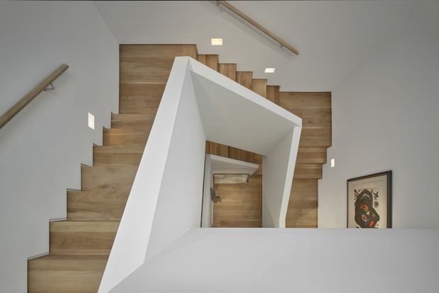 Interior view looking across a modern stairwell with a sculptural white angular balustrade and timber-clad stair treads and walls. White walls, recessed ceiling lights, timber handrails, and framed artwork contribute to a clean, contemporary architectural composition.