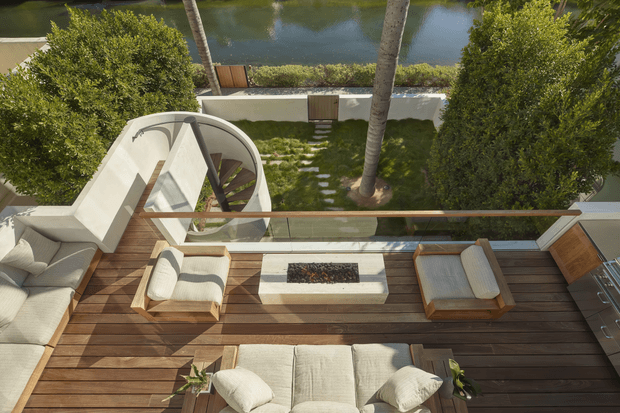 Elevated view of a refined outdoor terrace overlooking a landscaped courtyard and water feature. The terrace is furnished with cream upholstered lounge seating, a pair of sun loungers, and a central rectangular built-in fire pit set into a dark timber deck. A curved white architectural stair or sculptural structure connects to the garden below, while glass balustrading and timber detailing define the terrace edge. The overall design is warm, contemporary, and resort-like.