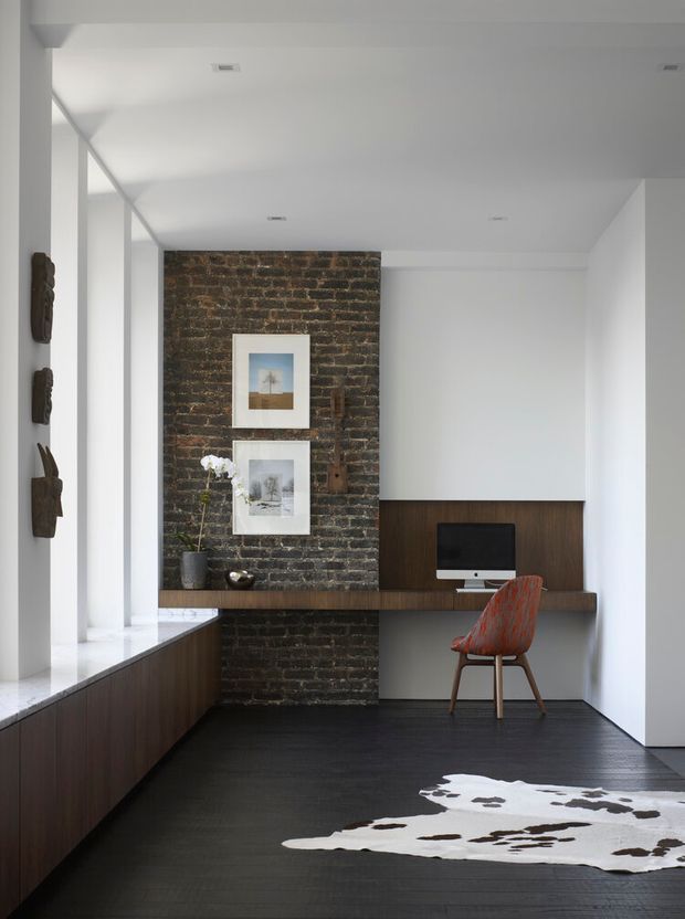A refined home office with a compact desk built into dark timber cabinetry, set against an exposed dark brick feature wall. The room has white walls and ceiling, a black timber floor, large side glazing, minimalist recessed lighting, a desktop computer, and a distinctive rust-coloured upholstered chair. A cowhide rug adds contrast in the foreground.
