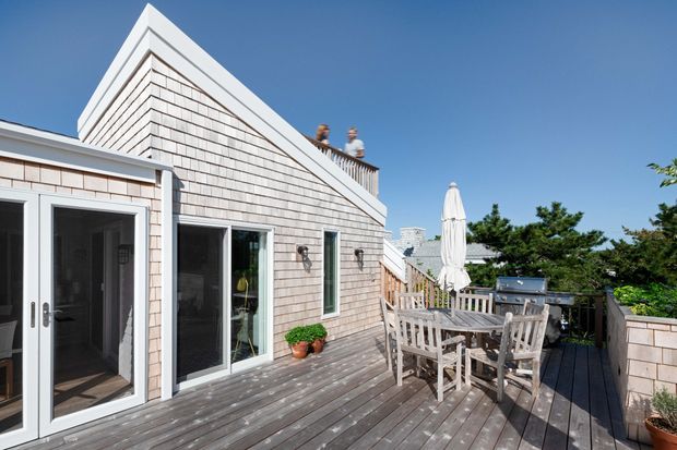 A sunny rooftop or elevated timber deck terrace beside a coastal-style shingle-clad building. The space includes weathered grey decking, a timber outdoor dining table and chairs, a built-in barbecue, a closed umbrella, planter pots, and white-framed glazed doors and windows. The gabled facade has pale shingle cladding with crisp white trim, and a clear blue sky and surrounding greenery are visible beyond the terrace.