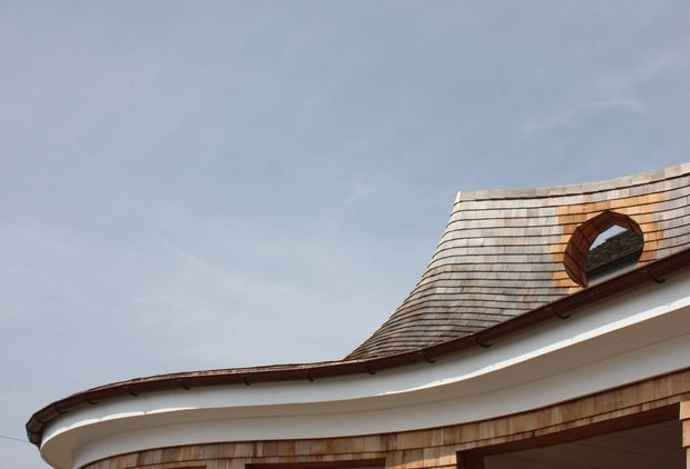 Close-up exterior view of a distinctive curved roofline and upper facade. The roof is clad in small overlapping timber shingles with a rounded dormer or oval window opening, while the facade below is white with brown timber trim and shingle detailing. The image focuses on the building envelope against a pale sky.
