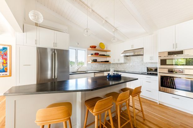 Bright, spacious white kitchen with a large dark island and four timber bar stools. The room features white shaker-style cabinetry, stainless-steel appliances, marble-look splashback, open timber shelving, pendant lights, and a white-lined pitched ceiling. Warm timber flooring and colourful ceramic accessories add contrast.