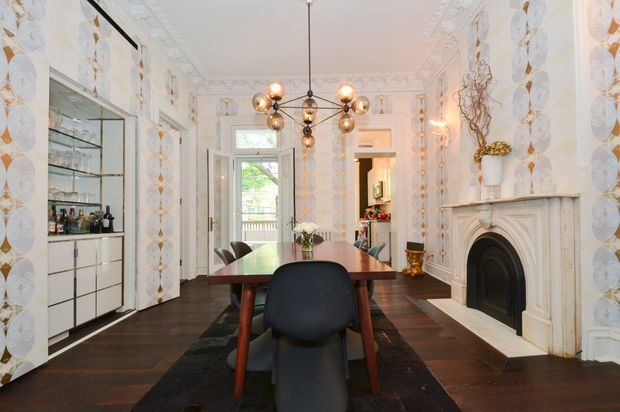 A refined formal dining room with a long dark timber dining table and sculptural black chairs, set against highly decorative white patterned wall coverings and ornate ceiling cornices. A marble or painted-stone fireplace with a dark arched firebox anchors the right wall, while a built-in bar and glass shelving occupy the left. A contemporary brass-and-glass globe chandelier hangs centrally, with French doors opening toward a balcony or exterior view. The room combines heritage architectural detailing with dramatic contemporary furnishings and lighting.