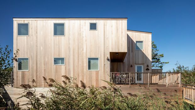 Exterior view of a contemporary multi-level residence clad in pale vertical timber boards. The rectilinear form includes a taller main volume, a lower projecting section, dark-framed rectangular windows, a recessed covered entry or balcony, and a timber deck with railing. Coastal vegetation is visible around the property under a clear blue sky.