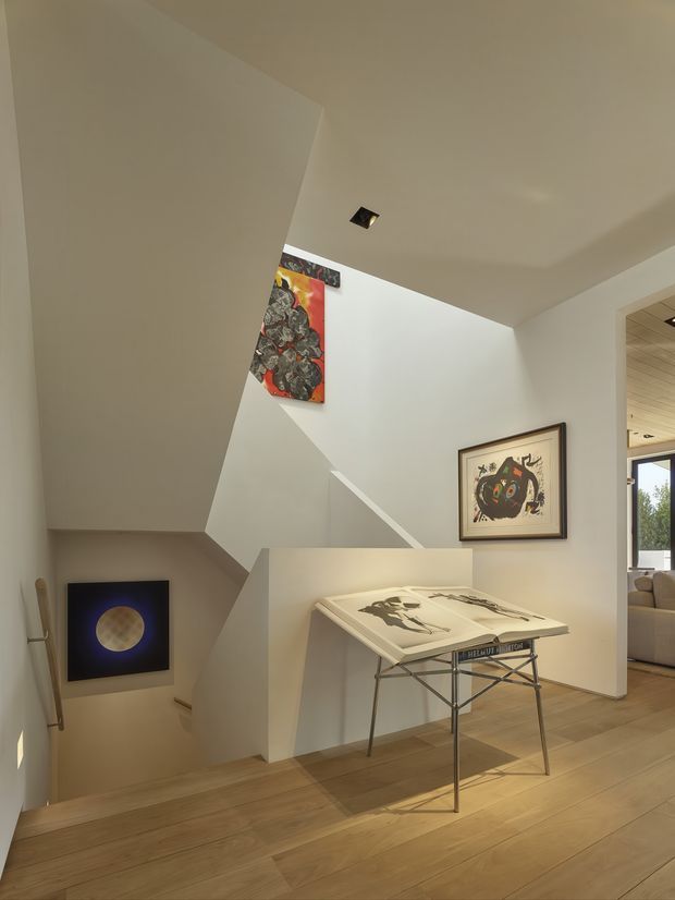 A contemporary residential stair landing with angular white walls and a sculptural open stairwell. Warm timber flooring contrasts with the white plaster surfaces, while framed artwork and a drafting or display table occupy the landing. A living area is partially visible through the opening at right, and a recessed ceiling spotlight provides focused illumination.
