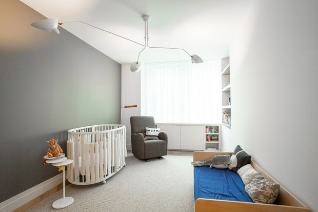 Bright, compact nursery and family room with a white round cot, grey upholstered armchair, built-in white shelving and cabinetry, a low timber-framed daybed or sofa, and a soft neutral carpet. A dark grey feature wall contrasts with the white ceiling, sheer curtains, and light joinery. Minimal sculptural pendant lighting and restrained furnishings create a calm, contemporary space.