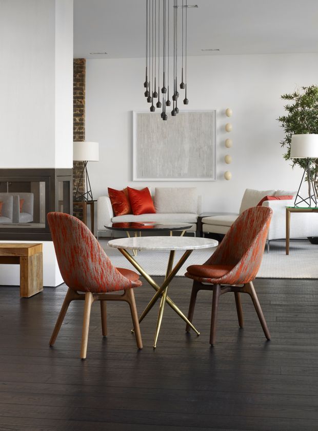 A refined contemporary living and dining area with dark timber flooring, a white fireplace wall, sculptural pendant lights, a pale upholstered sofa, and two distinctive rust-red lounge chairs around nested marble-topped tables. A partial exposed brick wall, large abstract artwork, indoor plants, and neutral furnishings add warmth and texture.