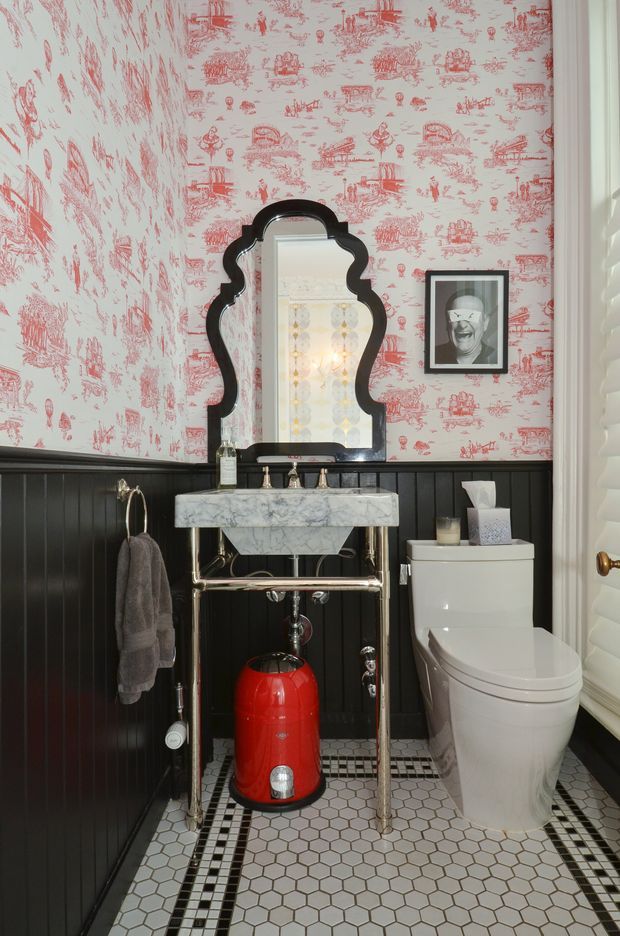 A compact, highly styled powder room with a white marble console basin on exposed polished metal legs, a sculptural black-framed mirror, white toilet, black beadboard wainscoting, and red-and-white toile-pattern wallpaper. The floor is finished in small white hexagonal tiles with black geometric border detailing. A bright red decorative object beneath the basin adds a bold accent.