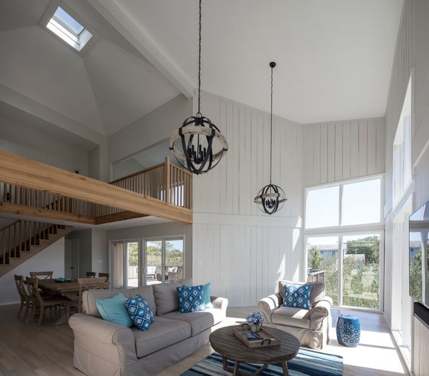 Bright double-height open-plan living space with a large grey sofa, armchairs, patterned blue cushions, a round timber coffee table and striped rug. White vertical wall panelling, expansive windows, skylight, and a timber mezzanine with balustrade and staircase define the architecture. Two dark sculptural pendant lights hang from the high ceiling, while a dining area is visible in the background.