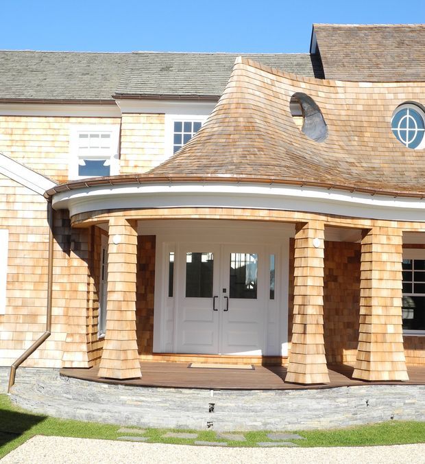 Exterior view of a distinctive shingle-clad building entrance, featuring a curved covered porch supported by tapered shingle-clad columns, white double entry doors, circular windows, and a highly sculptural curved roof form. The facade combines pale wood shingles with white trim and a low stone retaining wall.