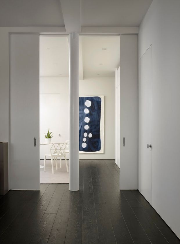 A minimalist interior hallway with dark timber flooring, white walls, sliding pocket doors, and a central round structural column. The hallway opens toward a bright dining area with a white table and geometric chairs, while a large blue abstract artwork provides a focal point at the far end. Clean recessed lighting and flush doors reinforce the contemporary, uncluttered design.