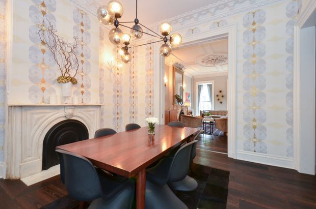 Elegant traditional dining room with a long polished timber dining table and sculptural dark upholstered chairs. The room features ornate white fireplace detailing, patterned decorative wallpaper, elaborate ceiling cornices and mouldings, dark timber flooring, and a contemporary clustered globe pendant light. An adjoining living area is visible through the wide opening.