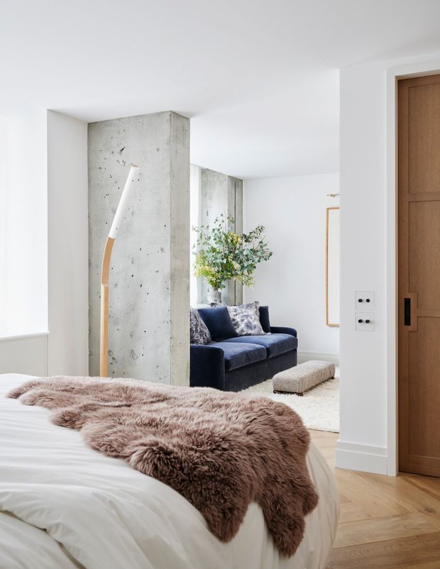 Bright, refined bedroom viewed toward an adjoining living area. The foreground features a white bed with a large taupe-brown sheepskin throw, pale timber parquet flooring, and a substantial timber door. A raw exposed-concrete structural wall divides the bedroom from the living space, where a dark navy sofa, patterned cushions, light rug, and leafy arrangement are visible. The palette is predominantly white and neutral with dark blue and natural timber accents.
