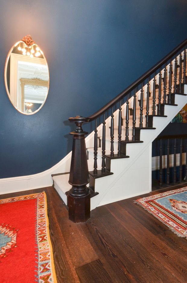 Interior view of a traditional staircase and entry hall. A dark stained timber staircase with turned balusters, a substantial newel post, and curved handrail rises along a deep navy-blue wall. White painted stair stringers and trim contrast with the dark timber floor. An ornate oval mirror hangs above the stair base, and richly patterned red and multicoloured runners frame the hardwood flooring.