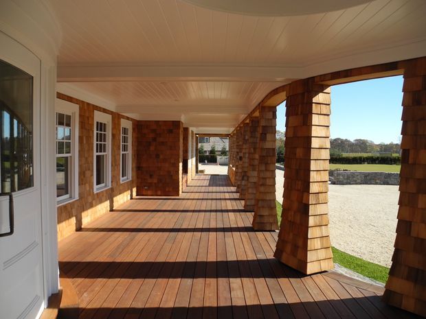 A long covered exterior veranda or porch with warm timber decking, shingle-clad walls and substantial shingle-wrapped columns. White-framed multi-pane windows and a white entry door line one side, while the open side overlooks a rural property. The veranda has a white tongue-and-groove ceiling and strong sunlight creating long shadows across the deck.
