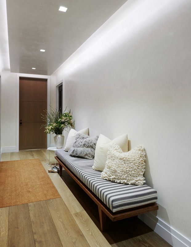 A narrow, minimalist hallway with white walls and a recessed ceiling featuring small square downlights. A long timber-framed bench with a black-and-white striped upholstered seat and layered cream and textured cushions runs along the right wall. Light timber flooring, a warm tan runner, a simple wooden door, a potted plant, and subtle decorative objects create a calm, refined entry passage.