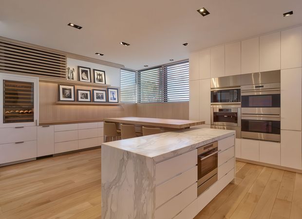 A refined open-plan kitchen and dining area with white full-height cabinetry, integrated stainless-steel appliances, a large marble-look island, timber flooring, and a timber dining table. Horizontal timber venetian blinds line the corner windows, while framed artwork and a built-in wine refrigerator add detail. Recessed spotlights provide even illumination.