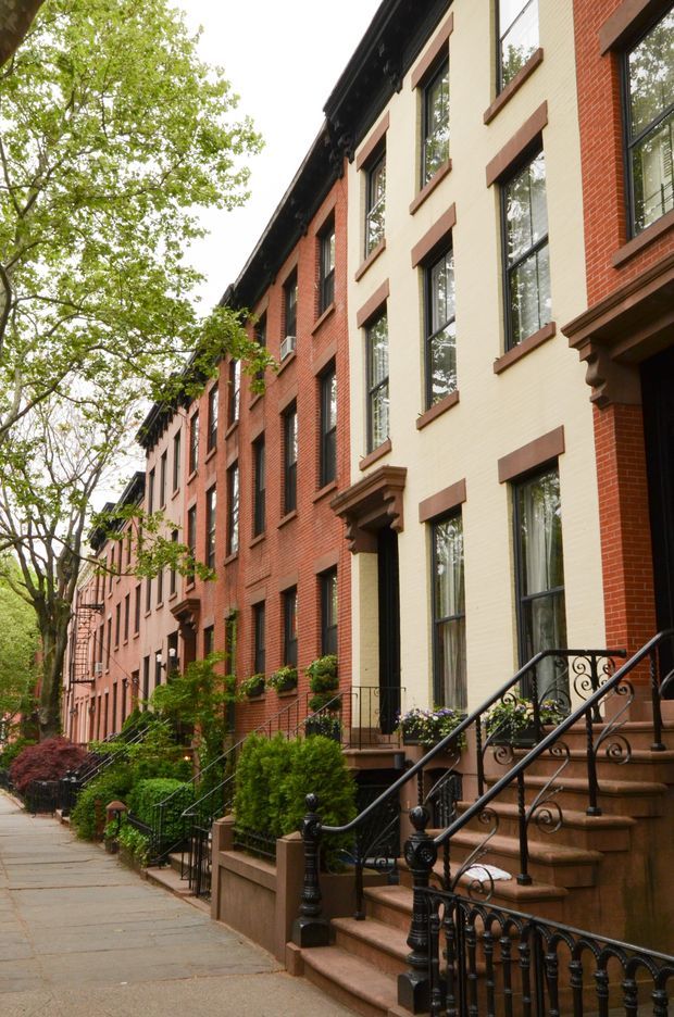 Street-level exterior view of a continuous row of narrow multi-storey attached townhouses. The façades combine red brick and pale painted brick, with tall dark-framed windows, projecting window surrounds, classical entry canopies, brownstone-like stoops, and ornate black metal railings. Mature street trees, planted front gardens, and a paved sidewalk line the residential urban streetscape.