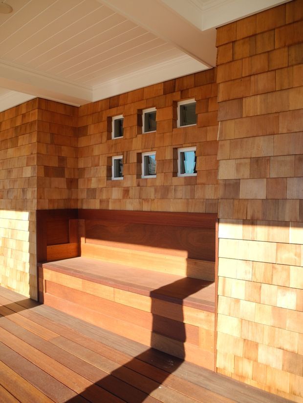 A covered timber-clad outdoor seating nook with built-in bench seating, surrounded by small square windows. The walls are clad in warm cedar shingles, with matching timber decking and a white-painted beadboard-style ceiling. Strong sunlight creates pronounced shadows across the bench and deck.