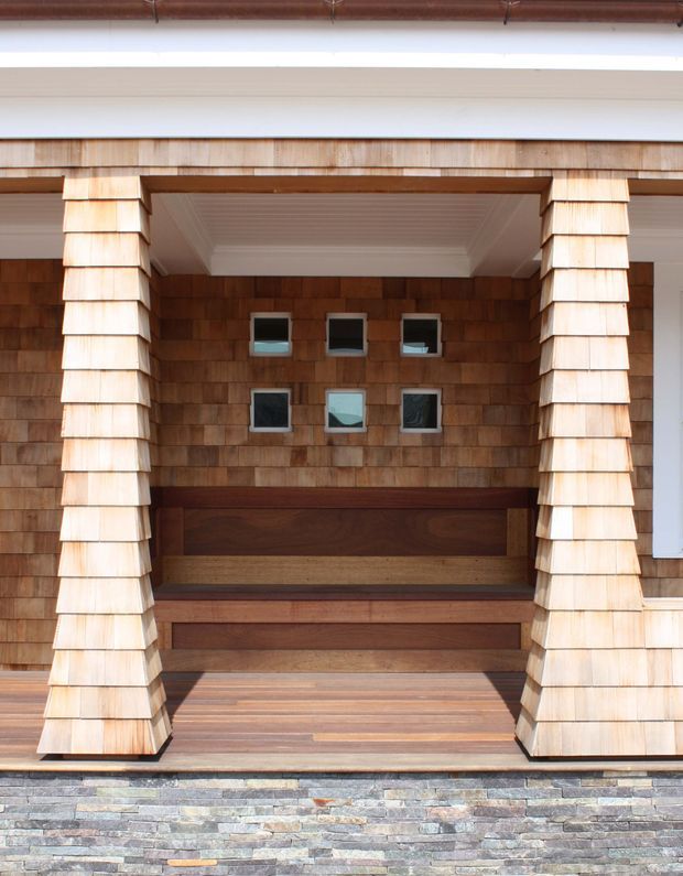 Close-up exterior view of a covered entry porch with warm cedar shingle cladding, chunky shingled columns, timber decking and built-in timber bench seating. A small grid of square windows punctuates the recessed wall, while layered stone veneer forms the base below. The composition highlights natural materials and strong crafted detailing.