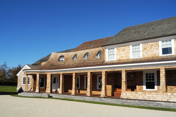 Exterior view of a large shingle-clad residential building with a long covered veranda, square timber posts, dormer-like upper windows, and a complex multi-section pitched roof. The warm weatherboard/shingle facade contrasts with white-trimmed windows and a broad pale gravel forecourt under a clear blue sky.