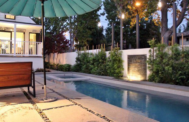 A landscaped residential pool courtyard photographed at dusk, with a long narrow swimming pool, pale stone paving, a lit textured feature wall and water blade, dense screening plants, and a covered outdoor seating area with a large green umbrella. A modern house with glazed doors and balcony railing is visible to the left.