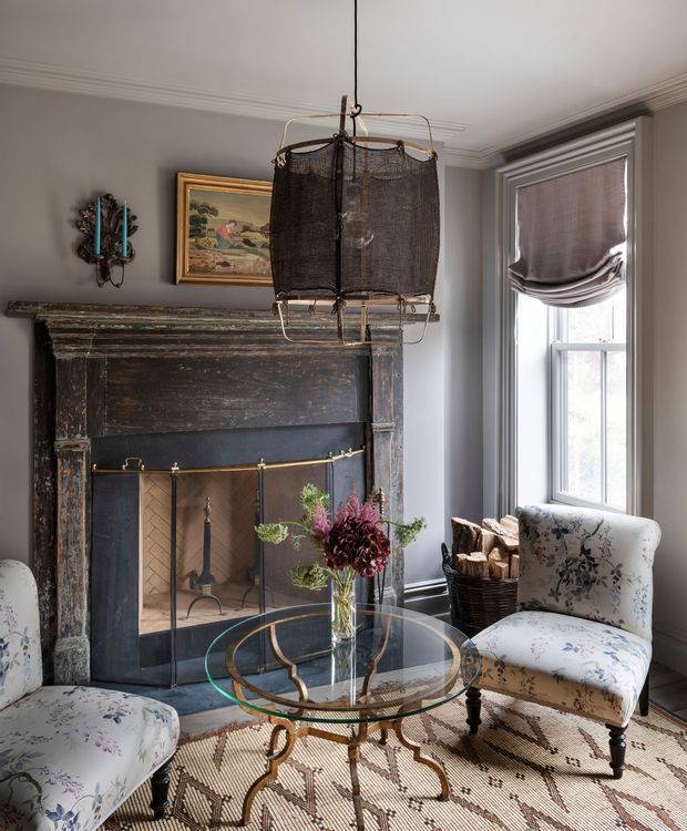 A refined traditional sitting room with a dark, aged timber fireplace surround, black firebox, and brass fireplace screen. Two floral upholstered armchairs frame a round glass-topped metal coffee table with a flower arrangement. A tall window with a fabric Roman shade provides natural light, while framed artwork, decorative wall lighting, a patterned rug, and ornate ceiling cornice complete the room.