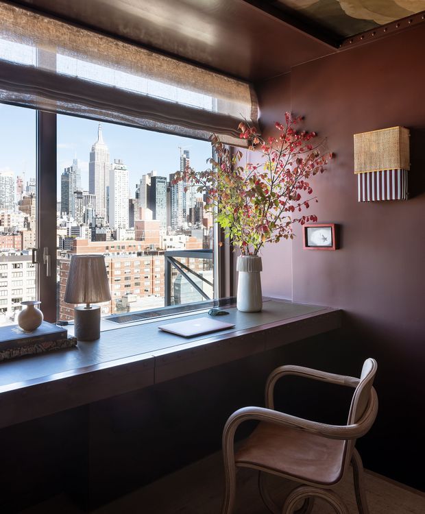 A compact, richly coloured home office or study nook set against floor-to-ceiling city glazing. A built-in dark desk runs along the window, with a laptop, table lamp, books, decorative vase and branch arrangement. A sculptural armchair faces the desk, while roller blinds filter bright daylight and provide a view toward a dense urban skyline featuring the Empire State Building. The room has deep burgundy walls and ceiling, dark built-in joinery, warm timber flooring, and layered decorative details.