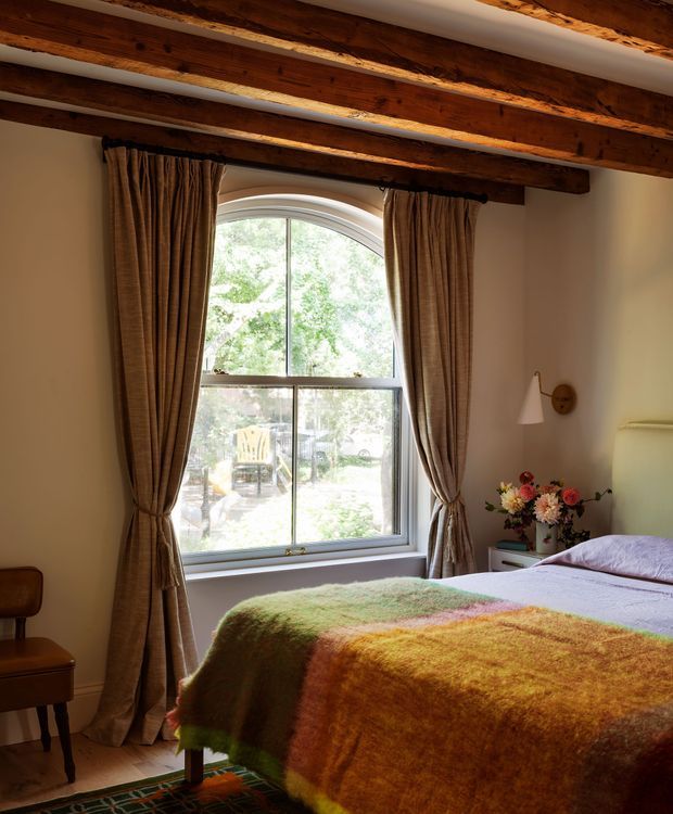 A warm, traditional bedroom with exposed dark timber ceiling beams, cream plaster walls, and a large arched-top window dressed with tied-back brown curtains. A bed with a colourful textured wool blanket occupies the foreground, accompanied by a small wooden bedside table, wall sconce, flowers, and a patterned rug. Natural daylight and the timber detailing create a cosy, rustic atmosphere.