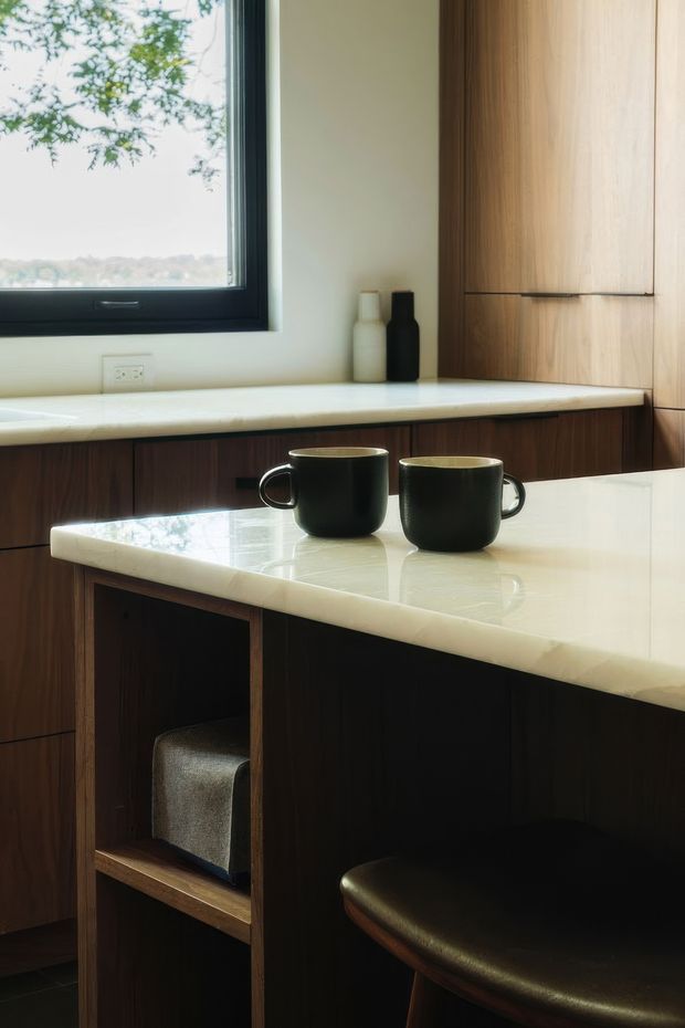 Close-up view of a refined kitchen featuring dark timber cabinetry, a pale stone waterfall-edge island benchtop, integrated seating, and a black-framed window overlooking greenery. Two dark ceramic mugs sit on the island, with simple bottles on the rear counter. The space has a warm, minimalist contemporary character with strong natural light.
