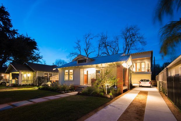 Twilight exterior view of a renovated single-storey suburban home with a blue metal gable roof, light horizontal cladding, brick feature wall, warm illuminated windows, landscaped front garden, and a long concrete driveway leading to a covered carport or garage area. A neighbouring bungalow is visible to the left.
