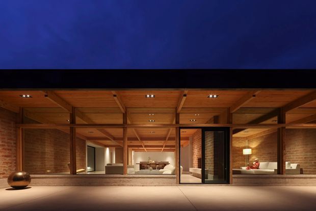 A dramatic dusk exterior view of a warm, timber-framed modern residence. The facade features extensive floor-to-ceiling glazing, exposed timber beams and soffits, and exposed brick walls. Interior living and dining areas are visible through the glass, with soft recessed lighting and warm furnishings. A broad paved terrace extends across the foreground beneath a dark night sky.